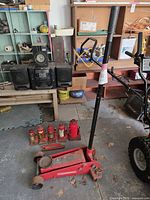 Hydraulic floor jack and four red bottle jacks placed on garage floor with some rust and used condition.