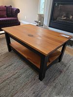 Coffee table angled view showing pine wood top and shelf with black frame and legs, minor wear visible on edges.