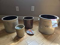Photo showing all five crocks of varying sizes, beige and brown glazed stoneware, alongside painted rock on hardwood floor.
