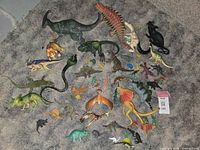 Lot of 50+ dinosaur and prehistoric creature figurines arranged on gray carpet, showing variety in size, color, and species.