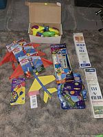 Overview photo showing all included toys arranged on a carpet, including boxed airplane kits, periscope kit, darts, kites, and water guns.
