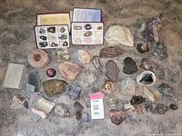 Overview of assorted rocks, minerals, and gemstone collection spread on surface with two labeled mineral specimen boxes, decorative stones, and a large amethyst cluster.