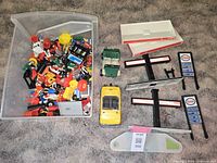 Overview showing a box of assorted Playmobil figures and station accessories alongside Esso branded canopy parts, sign boards, and two toy vehicles.