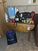 Full view of lot items arranged on a wooden table showing sleeping bag in carrying bag under table, lantern, radio, golf ball finders, rope, disposable cups, water jug, and other small items.