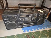 Full view of the Panasonic S-XBS stereo system showing main unit and attached speakers with visible dust and wear, placed on floor with foam mat under one speaker and cable on right side.