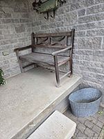 Wooden porch bench with carved backrest and slatted seat showing weathering, positioned on a concrete porch corner next to a brick wall