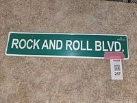 Front view of green metal street sign with white text 'ROCK AND ROLL BLVD.' lying on carpeted floor with seller tag nearby.