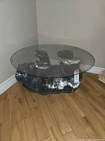 Front angled view of a black sculptural bear figure supporting a round clear glass coffee table top on wooden floor and light wall background.