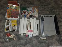 Photo of multiple crafting items spread on carpeted floor including rubber stamps grouped in plastic trays, various colorful plastic stencils with floral and animal designs, multiple white boxes containing stamps, a heavy-duty metal and plastic paper cutter with grid surface and handle, and pipe cleaners in a plastic bag.