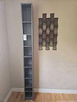 Tall narrow grey shelving unit standing upright next to wall showing height and compartments