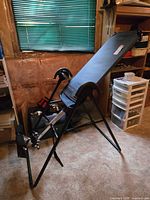 Teeter Hang Ups inversion table in a room with tan carpet and green blinds, showing side and front angles with padded backrest and metal frame.