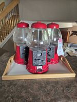 Three red metal bubblegum machines with clear globes placed on a rectangular wooden serving tray.