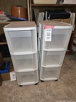 Front view of two white narrow plastic rolling carts each with three drawers, placed side by side on carpet floor.