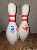 Two AMF Amflite II bowling pins, white with red stripes, showing blue ABC approval logo and red AMF logo. Used condition visible.