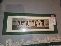 Two framed folk art prints each featuring black and white cows by Warren Kimble, with green wooden frames on the floor.