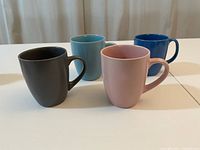 Four colourful ceramic mugs arranged in a row: black, light blue, pink, and dark blue.
