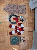 Top-down photo showing multiple Christmas decorations arranged on wood floor including fabric sack, two candy cane decorations, wreath, two candle holders, Christmas sign and Merry Christmas figurine