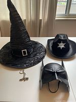 Photo of all four costume props: witch hat, sheriff hat, Batman mask, and cowboy boot keychain, laid out on white surface in front of window.