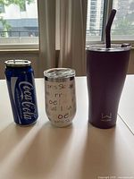 Photo of three drinkware items side by side, showing Coca-Cola can, holiday white tumbler, and purple tumbler.