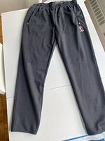 Front view of black Fila track pants laid flat on white background, showing zippered side pockets, elastic drawstring waist and Fila logo on left thigh.