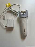 Front view of white Philips epilator with grey head and power button, alongside white charger with two-prong plug.
