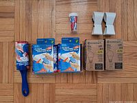 Photo showing the full collection of items laid out on wood flooring: lint roller with blue handle, two packs of lint roller refills, four mini glue tubes in a plastic container, two EcoSmart 60W LED bulbs in boxes, and two heavy-duty metal spring clips.
