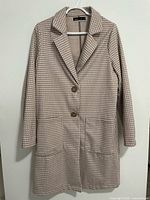 Full front view of the knee-length houndstooth coat hanging on a white hanger against a beige wall.