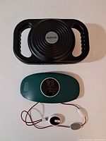 Black handheld vibration massager with ergonomic dual handles and circular massage surface; green EMS digital massager with digital readout and electrode pads connected by wires laid out below it on white surface.
