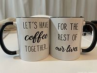 Two white coffee mugs side by side, both with black handles and rims, showing full phrases across the two mugs.