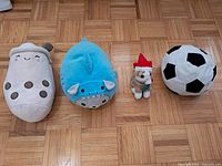 Four plush toys lined up on a wooden parquet floor: Boba Tea cup, blue shark, small Christmas cat, and a black and white soccer ball.