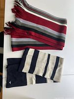 Photo of two scarves laid flat on a white surface, showing the red, white, grey, and black striped scarf with fringed ends and the black and cream striped scarf folded.