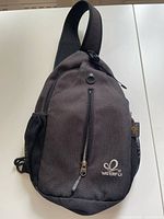 Front view of the Waterfly sling bag showing single shoulder strap, front zippered pocket, side mesh pockets, and brand logo