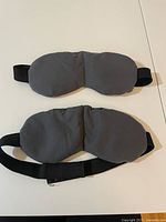 Top and front view of two gray weighted sleep masks laid flat showing front and back side along with adjustable black elastic straps.