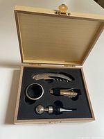 Open box showing wine accessory set inside black foam insert, containing corkscrew, bottle stopper, wine pourer, and drip ring