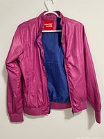 Pink raincoat with blue interior lining shown hanging on a white hanger, front view with zipper open.