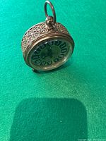 Clock angled slightly to show front Roman numeral dial, glass face and filigree metalwork on sides with loop handle on top.