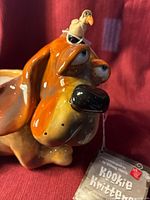 Close up front view of the ceramic dog candle holder showing its large eyes, black nose, and bird figurine on head with Kookie Kritters tag visible.