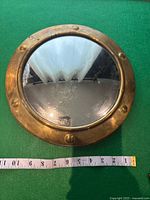 Front view of the antique brass convex porthole mirror on green fabric with measuring tape showing about 12 inches diameter.