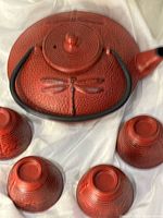 Red Japanese Tetsubin cast iron tea kettle with dragonfly detail on lid and side, shown with four matching red ridged tea cups arranged around it on white fabric.