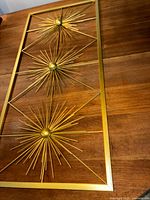 Gold metal wall art with three starburst designs on a wooden surface, captured from an angle showing the artwork's rectangular shape and detailed metal rod starbursts.