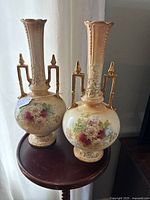Pair of tall hand-painted Royal Devon vases with gold gilt handles on a round wooden table near window light.