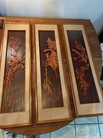 Full top-down view of three vertical rectangular carved wood panels showing birds and plant motifs, framed with beige border and gold trim on dark wood background.