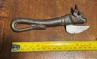Photo of an antique steel can opener shaped like a bull's head on the handle, shown next to a measuring tape indicating its size around 7 inches long.