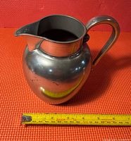 Front-right angled view of Armetale alloy pitcher on red background with a yellow measuring tape showing about 8 inches in height.