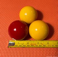 Three vintage billiard balls: two yellow and one dark red, placed on an orange surface with a measuring tape below showing each ball is about 2.3 inches in diameter.