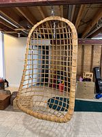Full view of light brown wicker hanging chair suspended by metal chain in a room with tiled floor and wooden ceiling.