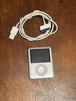 Apple iPod 8GB front showing screen and click wheel with white charging cable above it on wooden surface.