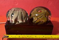 Front view of two Mdina glass paperweights with distinct swirl patterns on a wooden black stand, reference showing ruler for size scale.