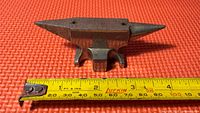 Top-down photo of the miniature metal anvil placed on a red textured surface with a yellow tape measure alongside showing length of approximately 4 inches