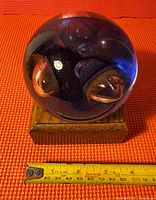 Glass sphere paperweight resting on an orange textured surface with a measuring tape showing it to be about 3 inches in diameter.